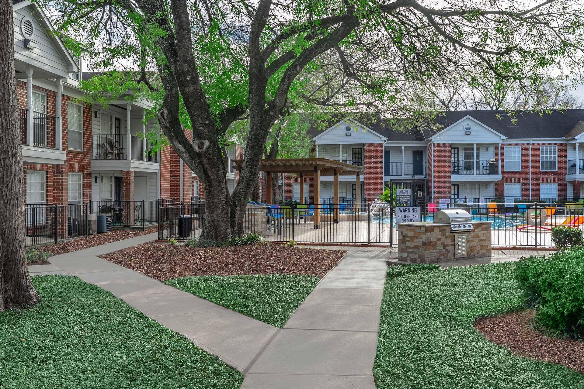 Apartment complex with pool and walkway. Brick buildings, trees, and a grill area.