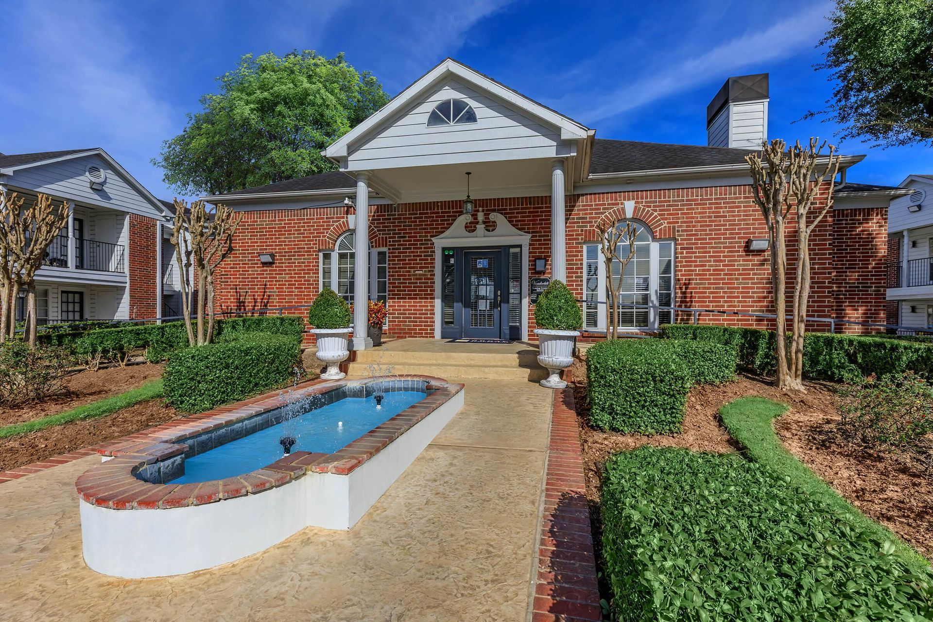Red brick building entrance with a fountain and trimmed hedges, blue sky.