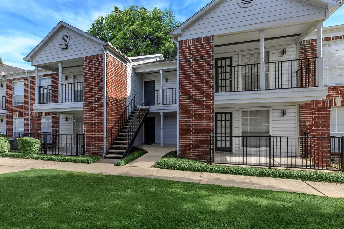 Apartment building with red brick accents, white siding, balconies, and black iron railings; green lawn in front.