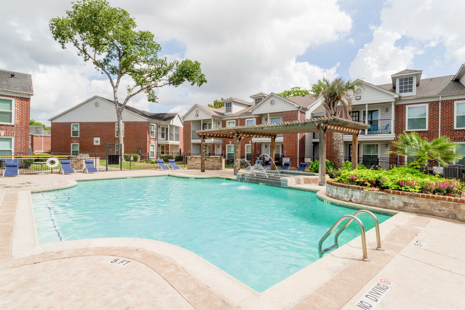 Red brick building entrance with a fountain and trimmed hedges, blue sky.