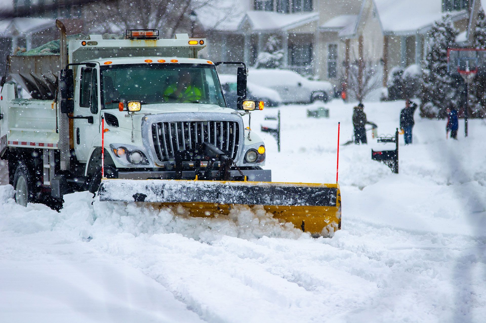 snow plow truck with yellow blade cleaning roads after winter storm with kids playing in the background