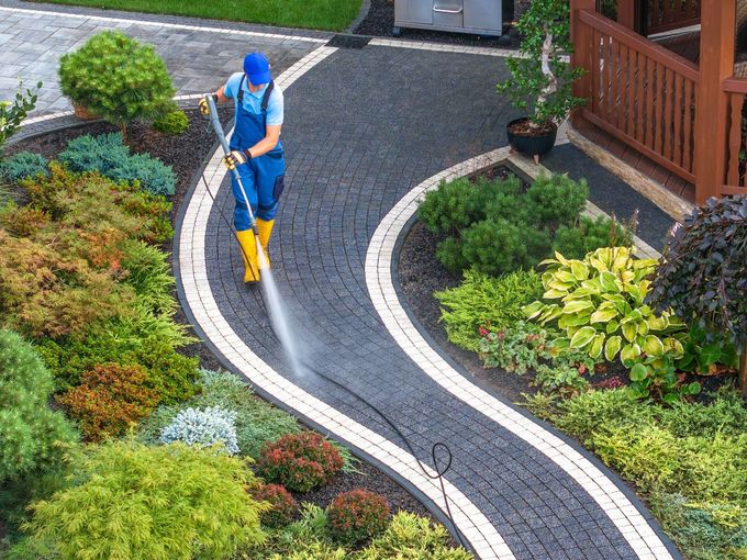 A man uses a high-pressure washer to clean a walkway in a garden.