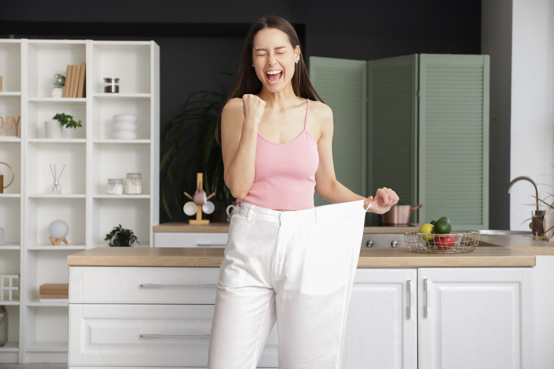 A joyful person in a pink top holds out the waist of their loose white pants, showing weight loss in a kitchen setting.