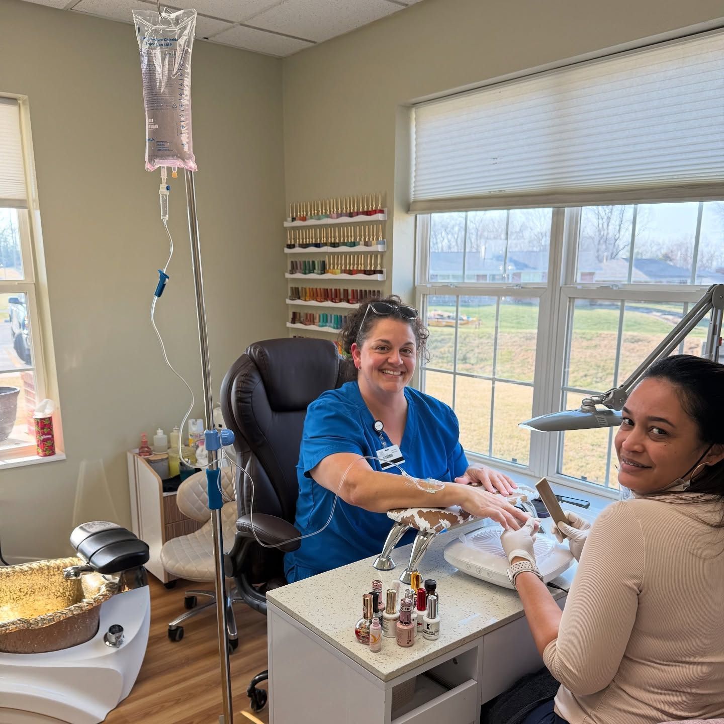 A person wearing medical scrubs with an IV drip attached sits at a nail salon desk while receiving a manicure.