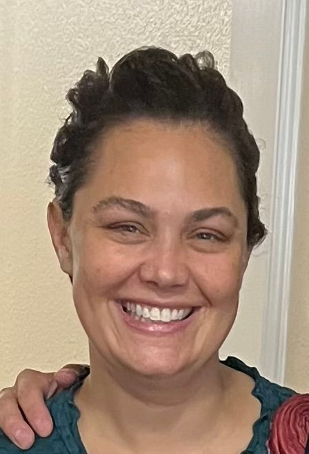 Woman with dark hair smiles, wearing teal top, arm around her shoulder.