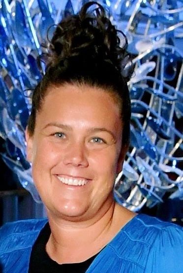 Woman with dark hair in a bun smiling, wearing a blue top, in front of a blue sculpture.