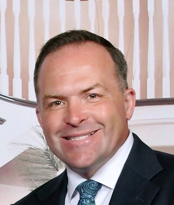 Man in suit smiles broadly, looking directly at the viewer. Background with light patterned wallpaper.