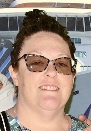 Woman wearing glasses smiles in front of a cruise ship, wearing a patterned shirt, and has dark hair.