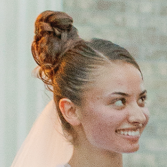 Woman with brown hair in a bun smiles, looking right; soft blush on cheeks, light background.