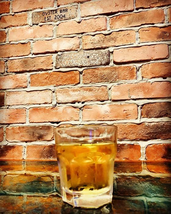 Glass of amber liquid with ice in front of a brick wall, possibly at a bar.