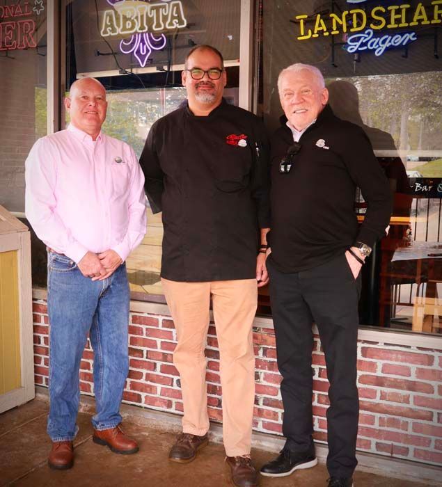 Three men standing outside a restaurant; chef in center, flanked by men in casual wear.