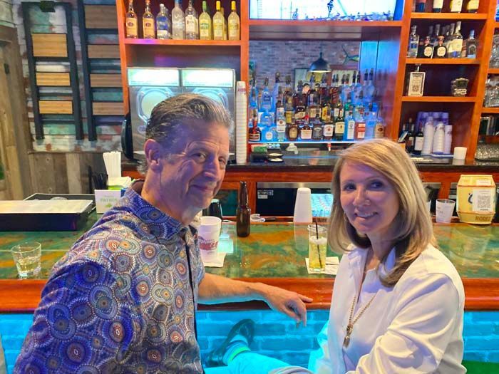 A man and woman sit at a bar. The man wears a patterned shirt, the woman a white one. Bar is stocked with bottles.