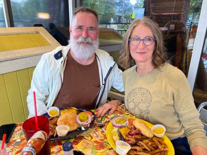 A couple smiles at a table with burgers and fries. They are seated in a diner-like setting.