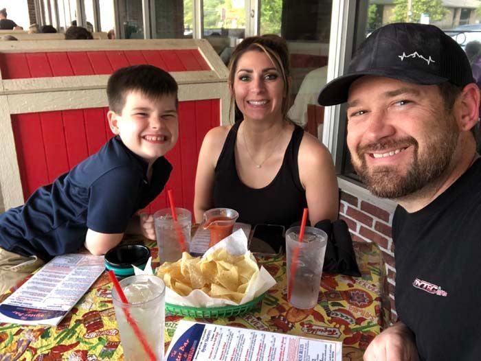 Family of three smiling at a table with drinks and chips at an outdoor restaurant.