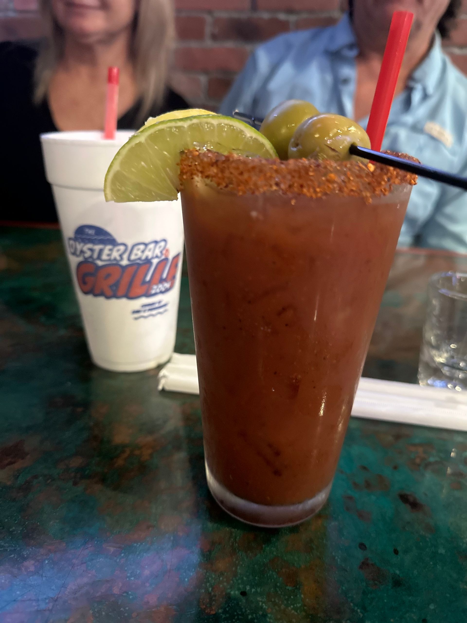 Bloody Mary drink with olive garnish and lime, on a table. Person in background.