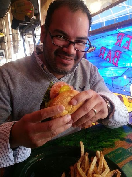 Man with glasses smiles, holding a sandwich with a side of fries at a table. 