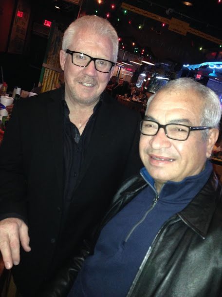 Two men smiling, wearing glasses, in a dimly lit bar. One in black blazer, the other in black leather jacket.