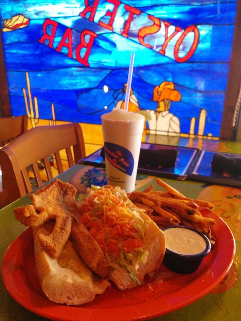 Plate of food at an oyster bar. A sandwich, fries, and drink on a table. Window behind it with an oyster bar sign.