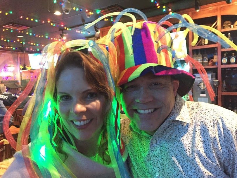 Two people smiling, wearing colorful, light-up party hats at a bar.