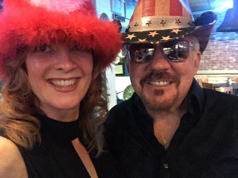 Woman in a red fluffy hat smiles next to a man in a cowboy hat with American flag stars.