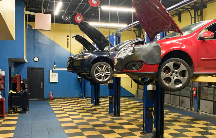 Two cars with open hoods elevated on hydraulic lifts inside an auto repair shop with a yellow-and-black tiled floor. | Airpark Auto Pros