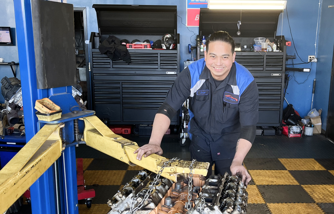 A smiling mechanic in a shop uniform works on a vehicle engine component in a garage with large tool cabinets. | Airpark Auto Pros