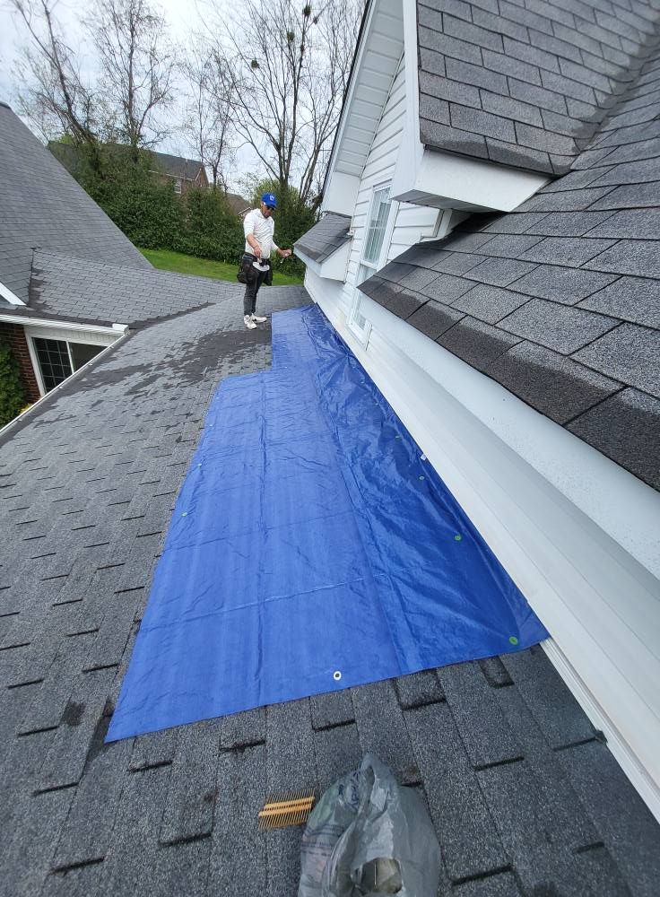 Person on a roof covered in a blue tarp. Shingled roof next to a white house. Cloudy day.