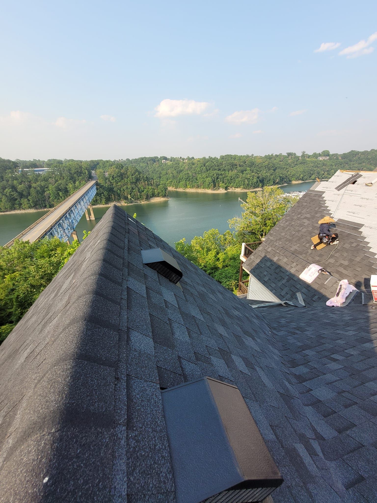 Damaged house roof with a river, bridge, and trees in the background under a blue sky.