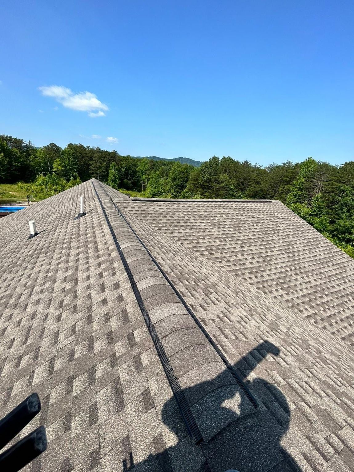Rooftop with gray shingles on a sunny day. Trees in the background, blue sky overhead.