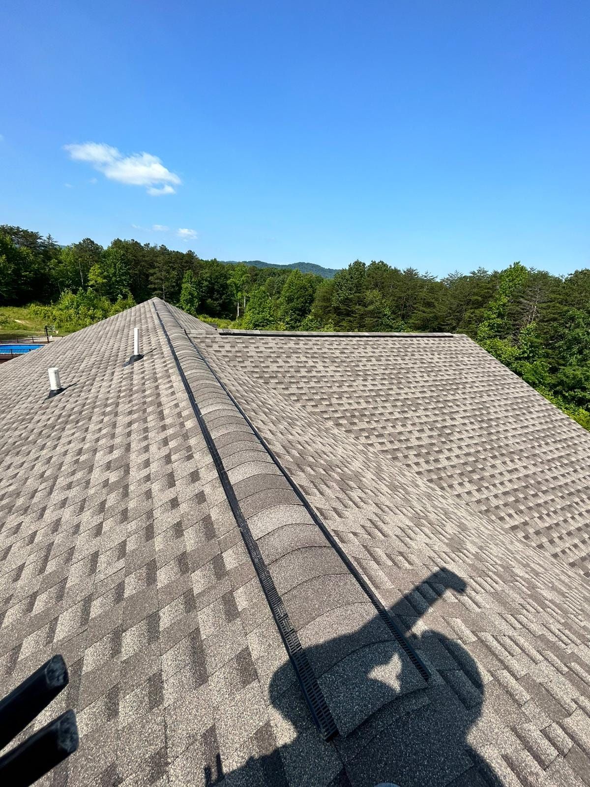 Rooftop with gray shingles on a sunny day. Trees in the background, blue sky overhead.