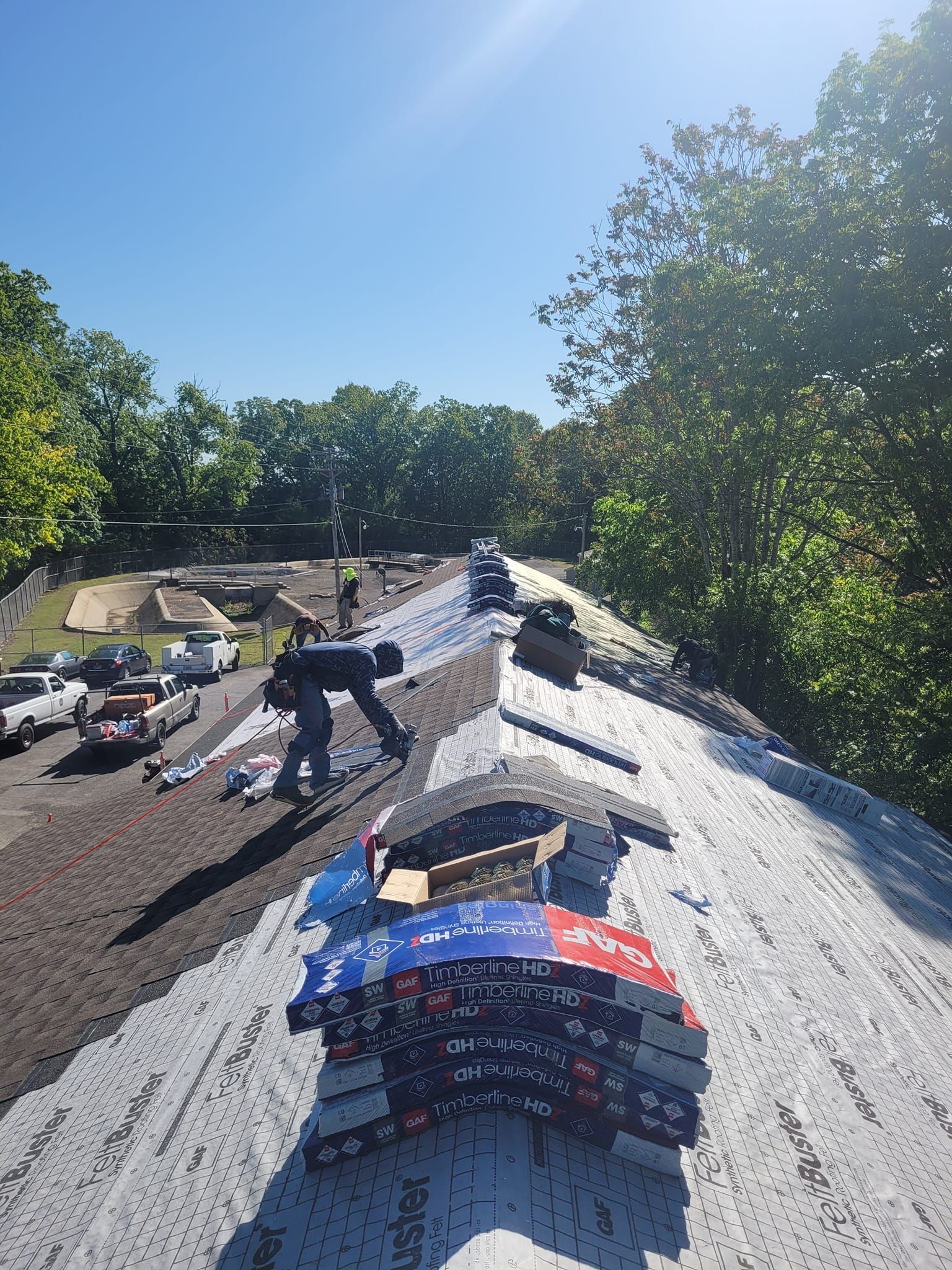 Roofers installing shingles on a roof under a bright blue sky. Several stacks of shingles are visible.