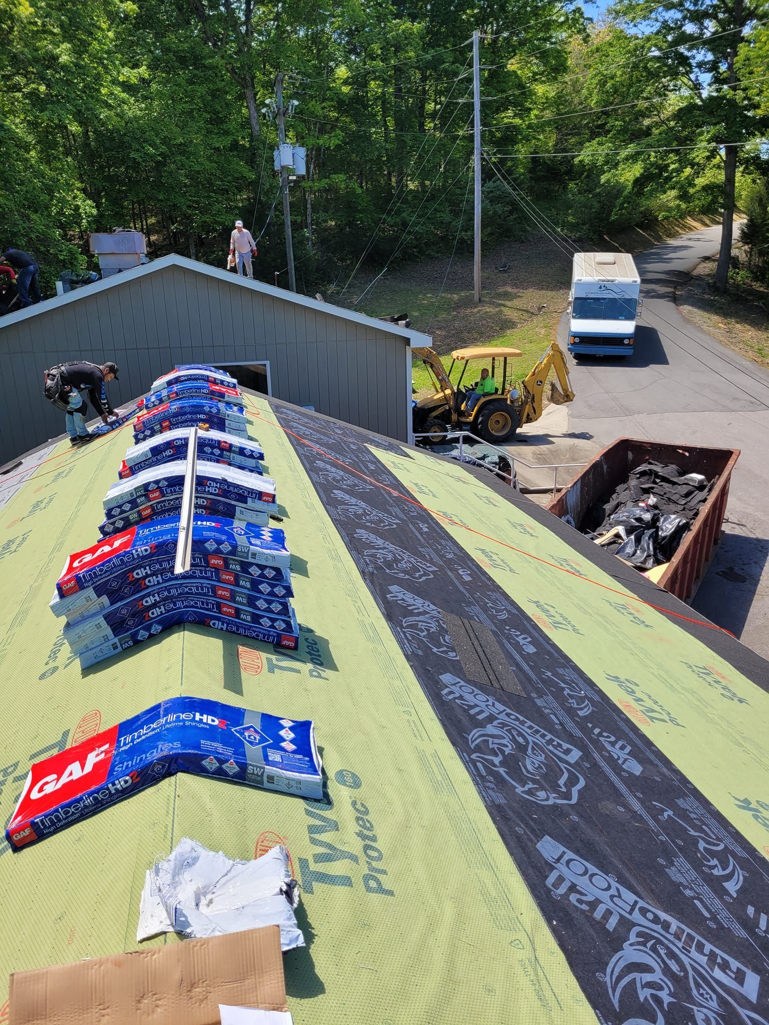 Roof with stacked bundles, black underlayment, and a dumpster. A truck and yellow loader are in the background.