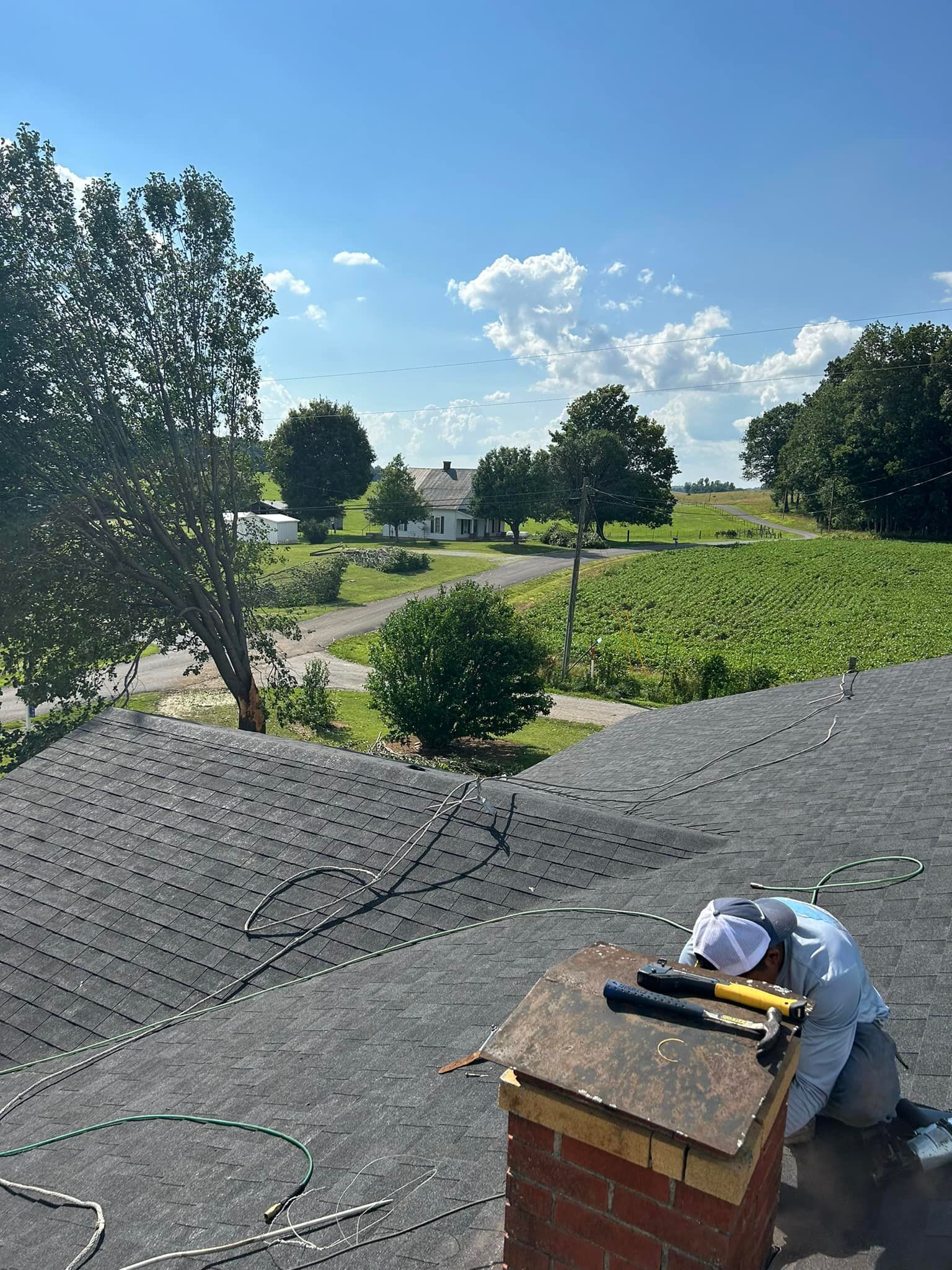 Roofer kneeling on roof near brick chimney, using tool. Green trees and fields in background. Blue sky, sunny day.