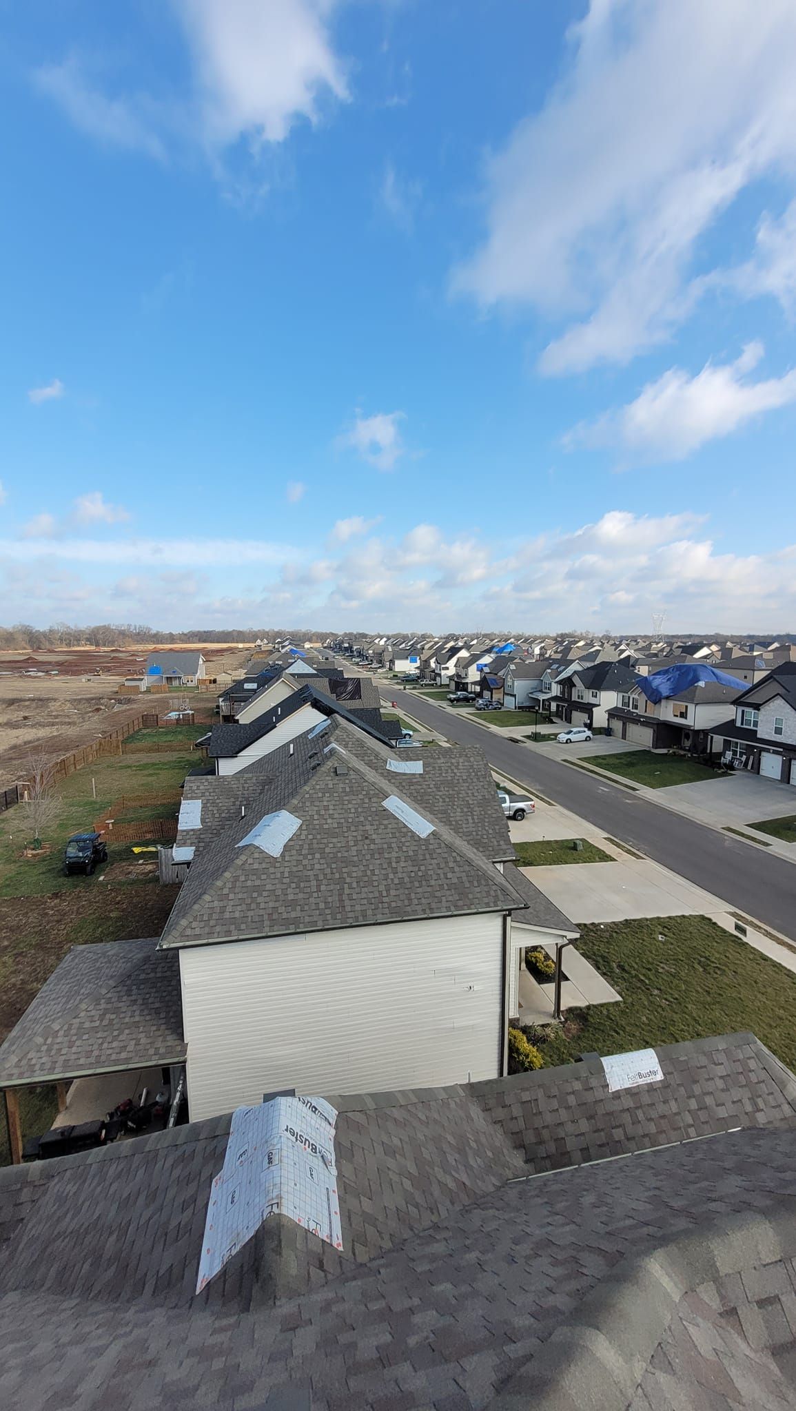Rooftop view of a neighborhood with rows of houses, gray roofs, and blue sky with clouds.