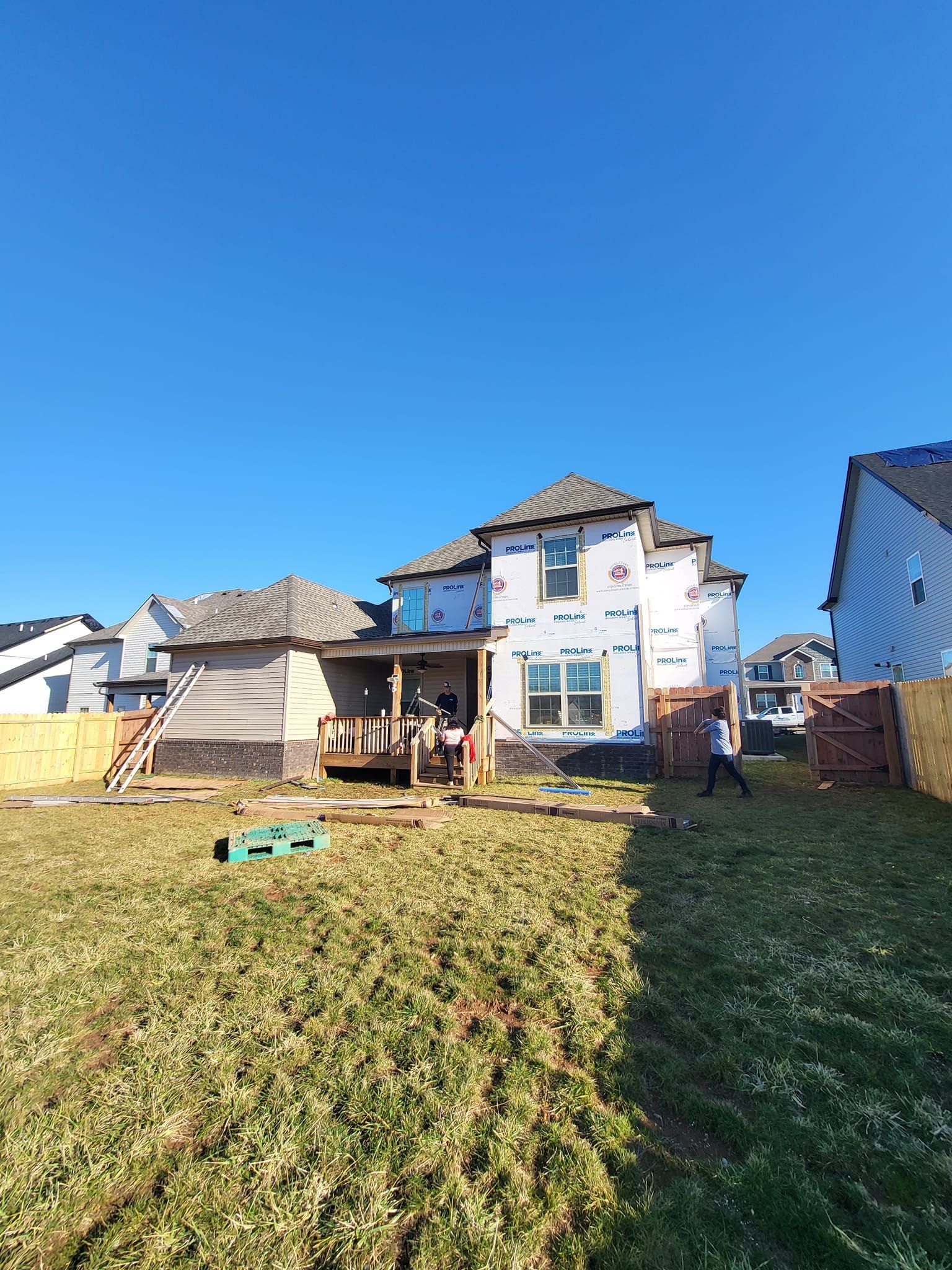 Back of a house under construction; blue wrap, unfinished siding, blue sky, and a grassy yard.