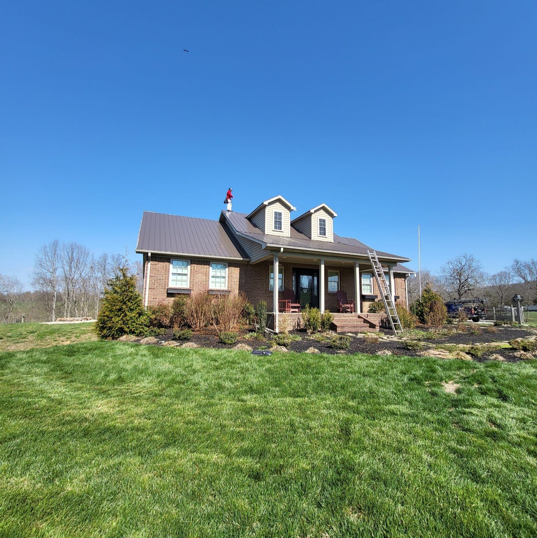 Brick house with porch, dormers, and dark roof on a grassy hill under a blue sky. Ladder leans against the house.