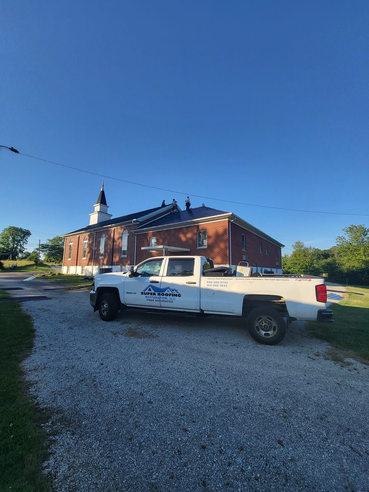 White truck parked near brick building with workers on the roof under a blue sky.