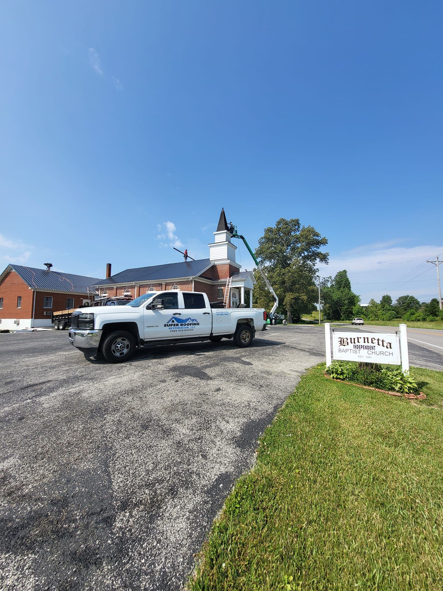 White truck parked on gravel, near a building with a white steeple. A crane extends toward the roof, sunny day.