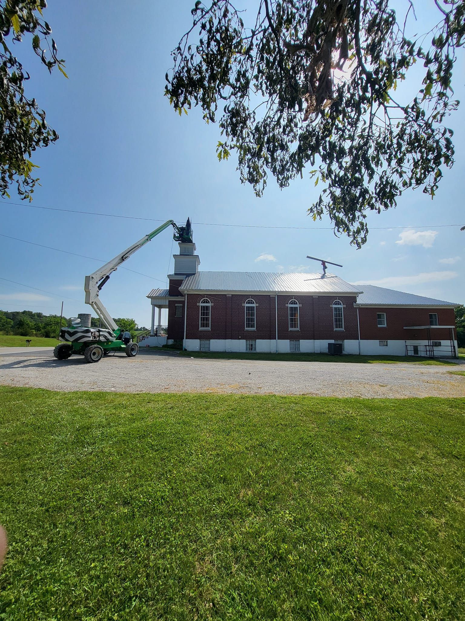 Brick building being worked on by a lift on a sunny day. Green grass and trees frame the scene.