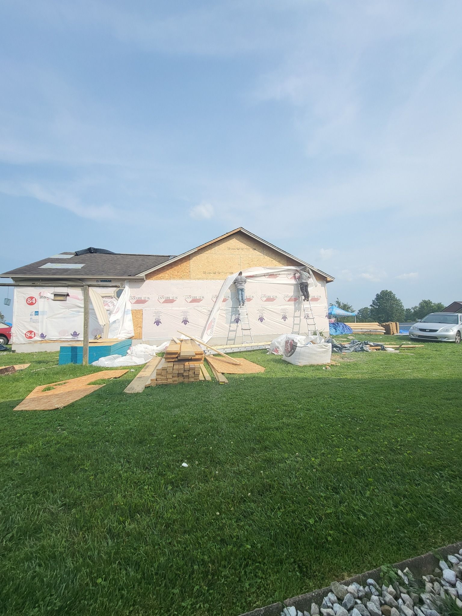 A house under construction; siding is being installed. Boards and materials sit on the lawn. Bright, sunny day.