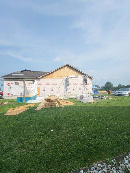 Construction of a house: plywood walls, roofing, and materials on the grass under a blue sky.
