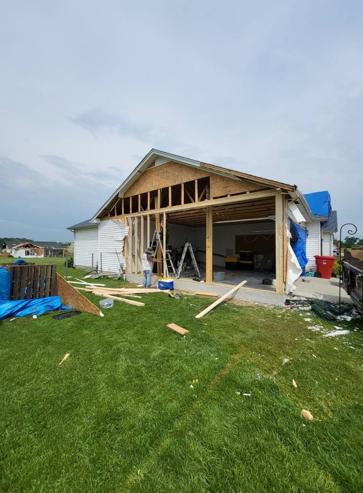 Garage undergoing construction, open-framed walls. Blue tarp on roof, tools and debris on lawn. Overcast sky.