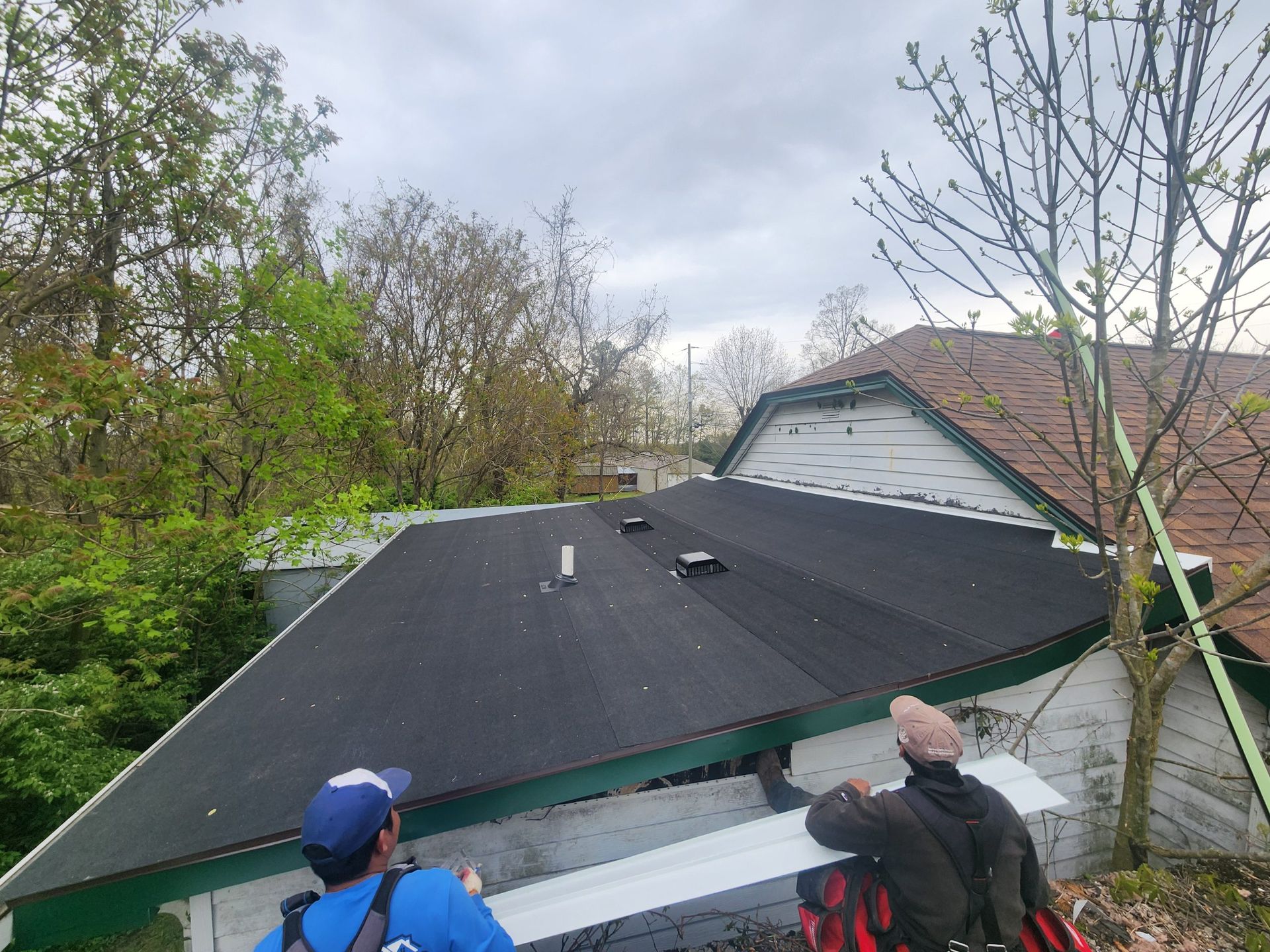 Two workers installing roofing material on a low-sloped roof with trees in the background under a cloudy sky.