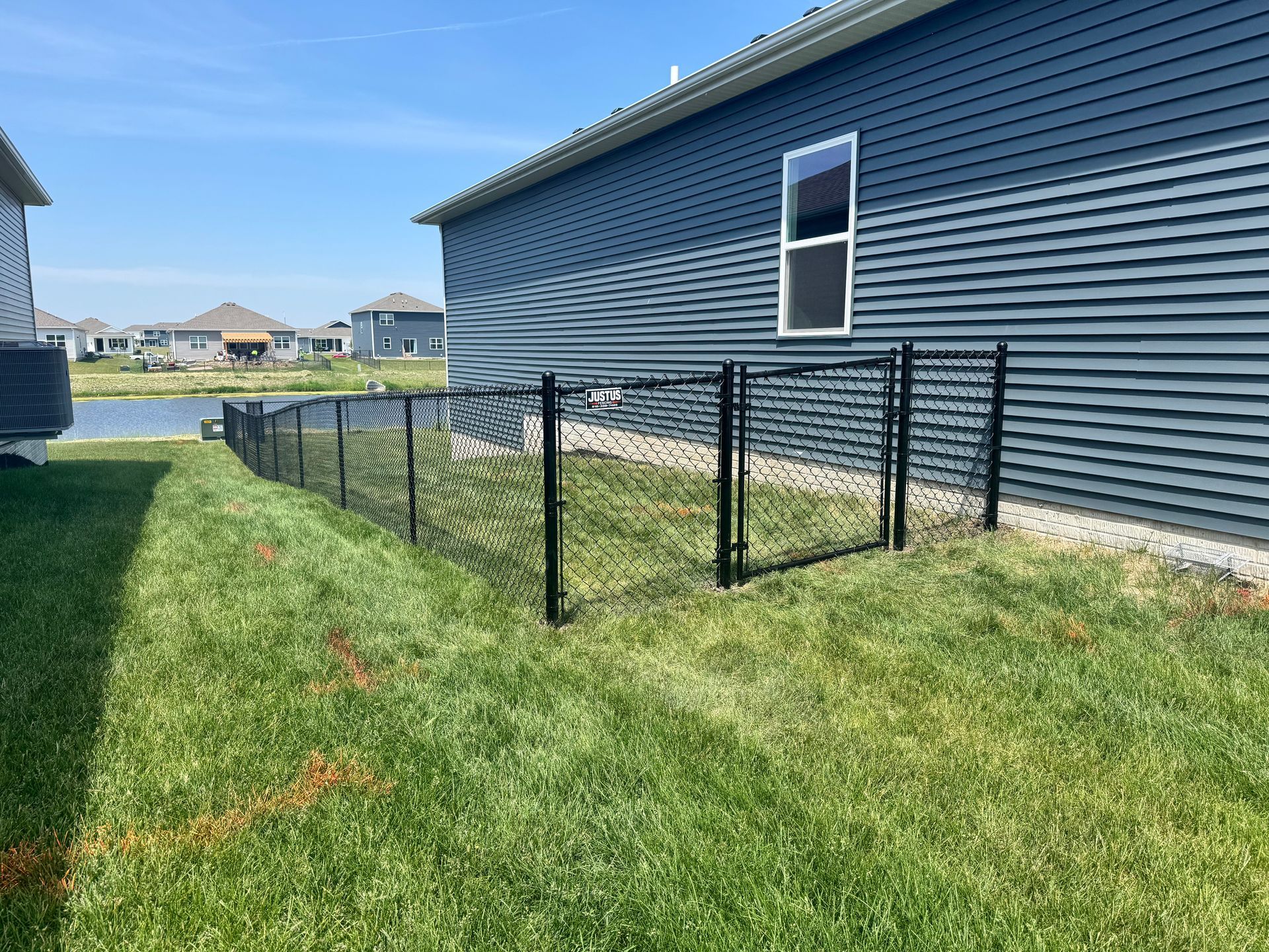 Black chain-link fence encloses a green lawn between two-story houses on a sunny day.