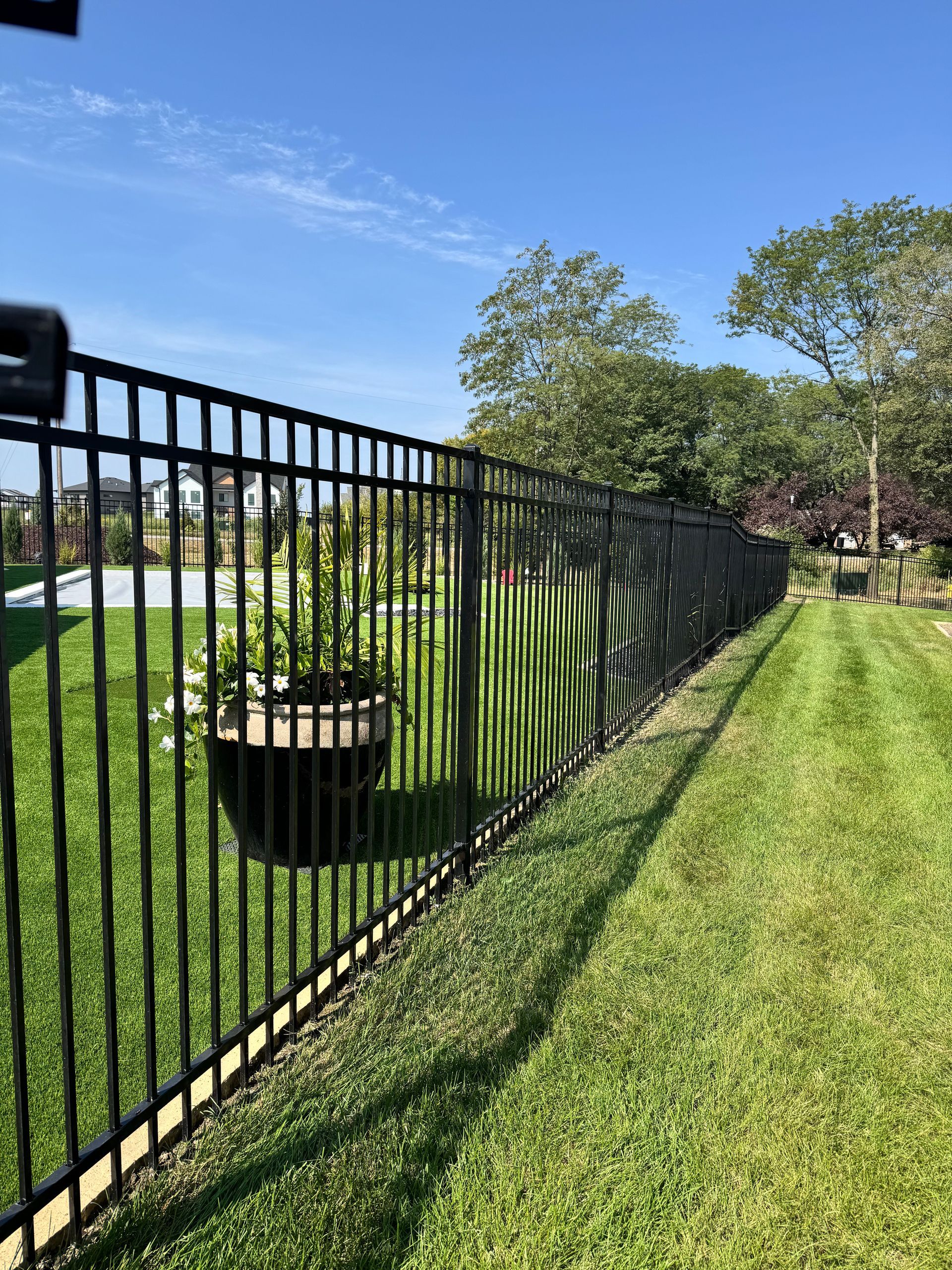 Black fence curves along a grassy yard, with trees on the left and a cloudy sky in the background.
