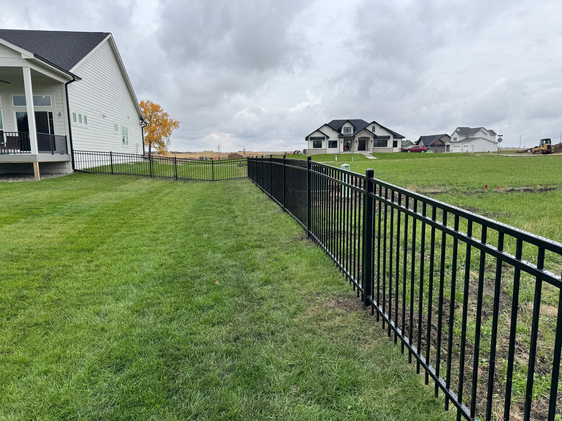 Black fence curves along a grassy yard, with trees on the left and a cloudy sky in the background.