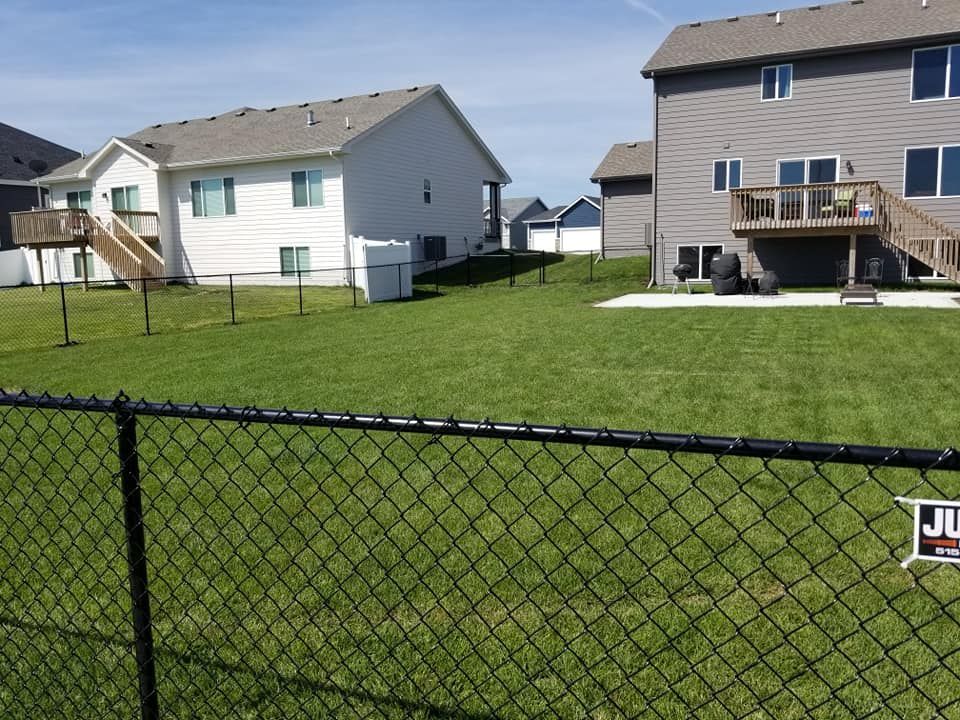 Black chain-link fence encloses a green lawn between two-story houses on a sunny day.