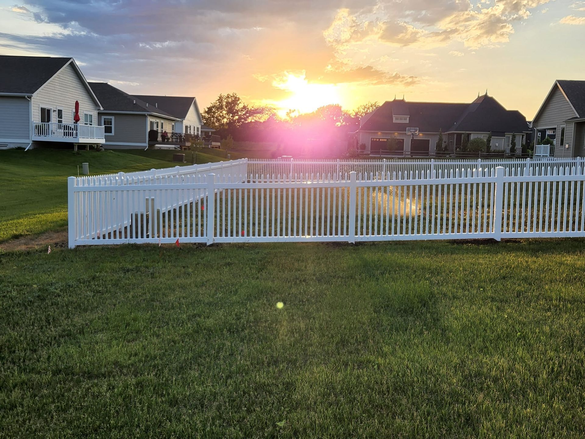 White picket fence around a pool, with a sunset over residential homes.