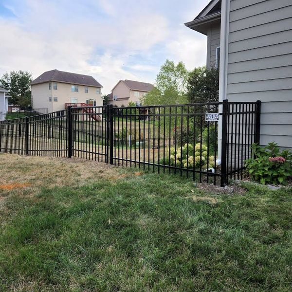 Black metal fence around a yard with a house, grass, and neighboring houses in the background.
