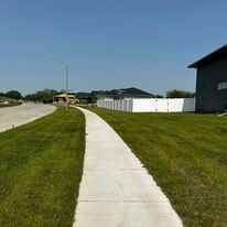 Concrete sidewalk through green grass, houses, and bright blue sky.